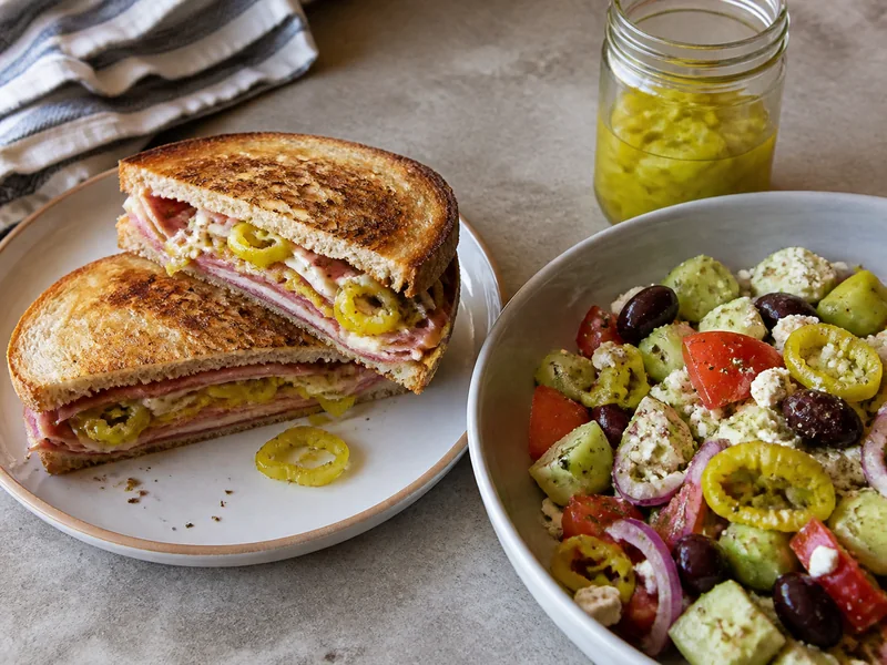 Banana pepper rings in a sandwich beside pepperoncini in Greek salad and a jar of brine