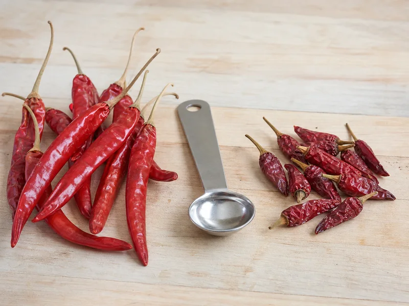 Long smooth De Arbol pods beside shorter wrinkled Tien Tsin chiles with a spoon for scale