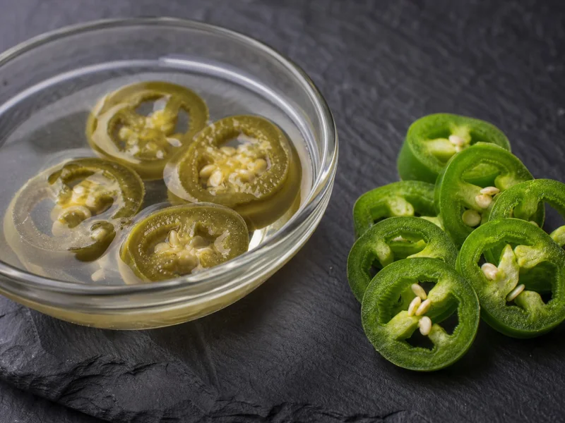 Pickled jalapeno rings in brine beside crisp fresh jalapeno slices