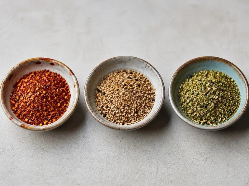 Three bowls of shichimi with red chile-led, sesame-led, and green herb-led colors and textures
