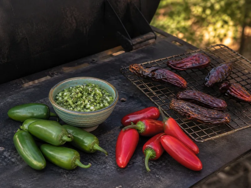 Fresh green jalapenos and salsa beside ripe red jalapenos and smoked dried pods on a rack