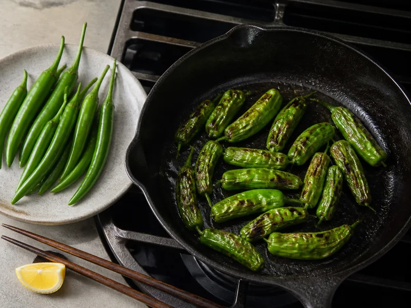 Blistered Shishito peppers in a pan beside uncooked long Fushimi peppers