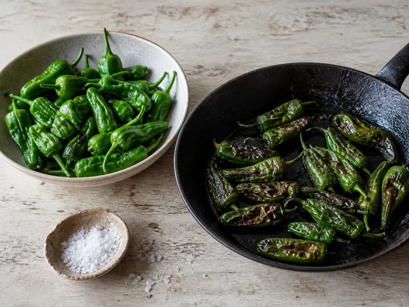 Fresh and blistered green Padrón peppers beside a skillet and coarse salt