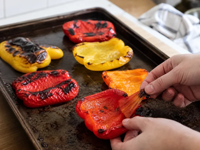 Hands peeling blistered skin from a roasted red bell pepper beside roasted yellow, orange, and red halves