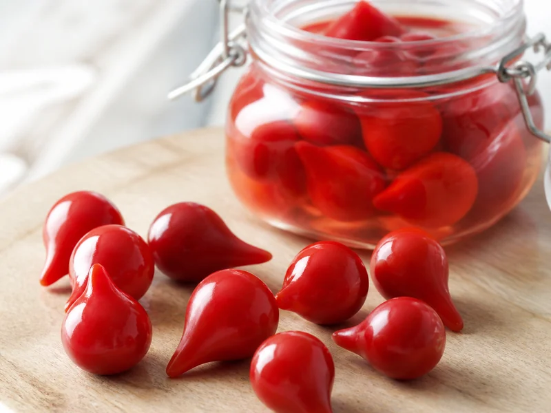 Small bright red biquinho peppers showing the pointed beak tip that gives the pepper its name, scattered on a board beside an open jar of brine