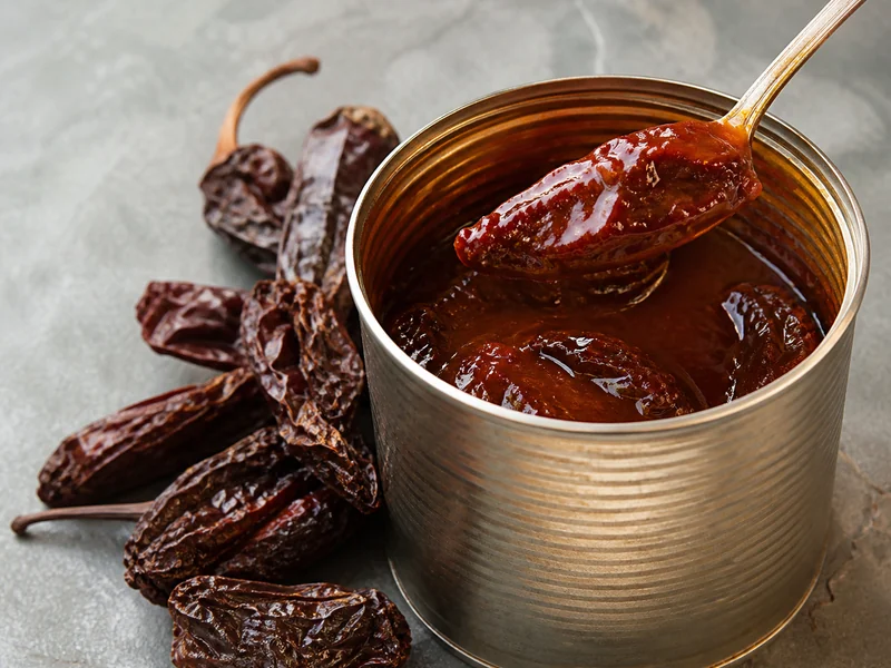 An opened tin of chipotles in dark adobo sauce with a spoon lifting one, beside dried whole chipotle pods showing wrinkled leathery skin