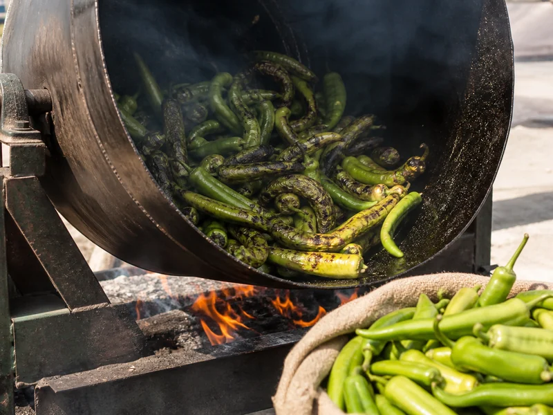Green Hatch chiles blistering in a rotating drum roaster over open flame, with a sack of fresh pods beside