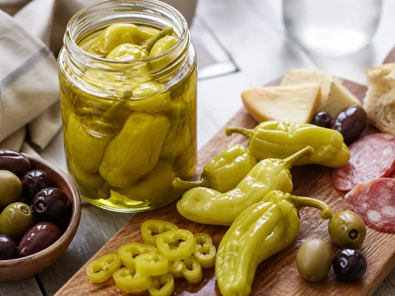 Pale yellow-green pickled pepperoncini in an open jar of brine, with drained pods and sliced rings on an antipasto board