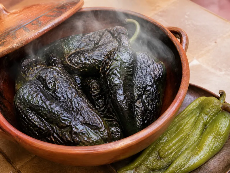 Charred poblanos steaming under a lid in an earthenware bowl, with one peeled pod beside showing pale green flesh