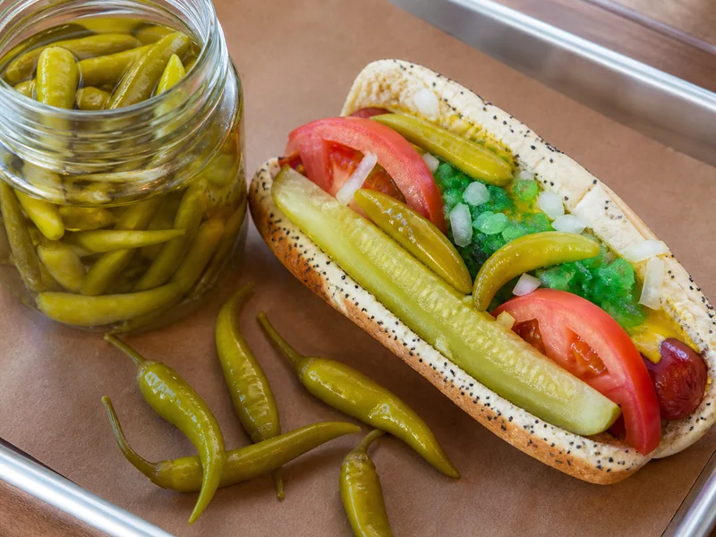 Small green pickled sport peppers in a jar beside a Chicago-style hot dog