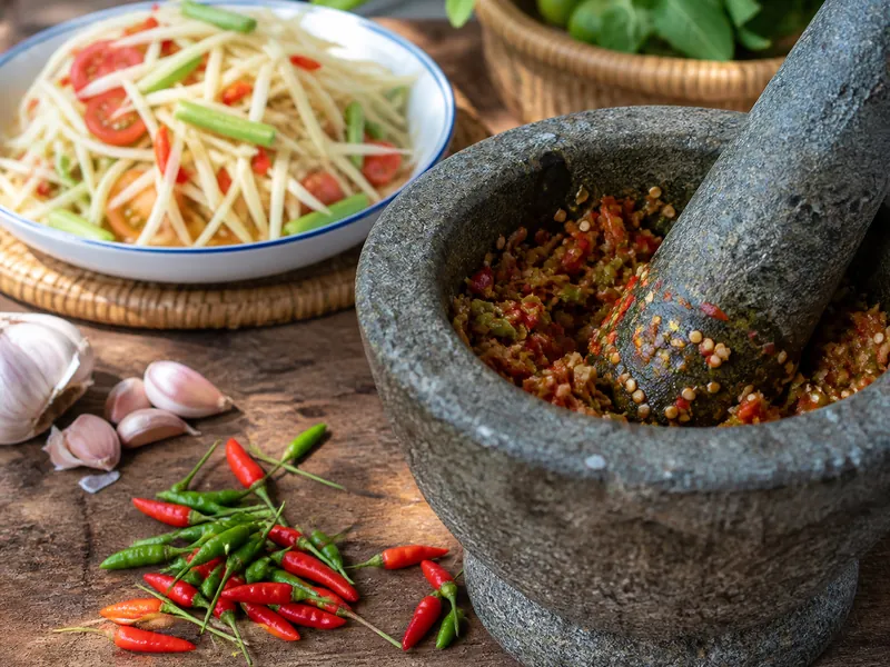 Small red and green Thai chilies whole on the table and pounded in a granite mortar beside green papaya salad