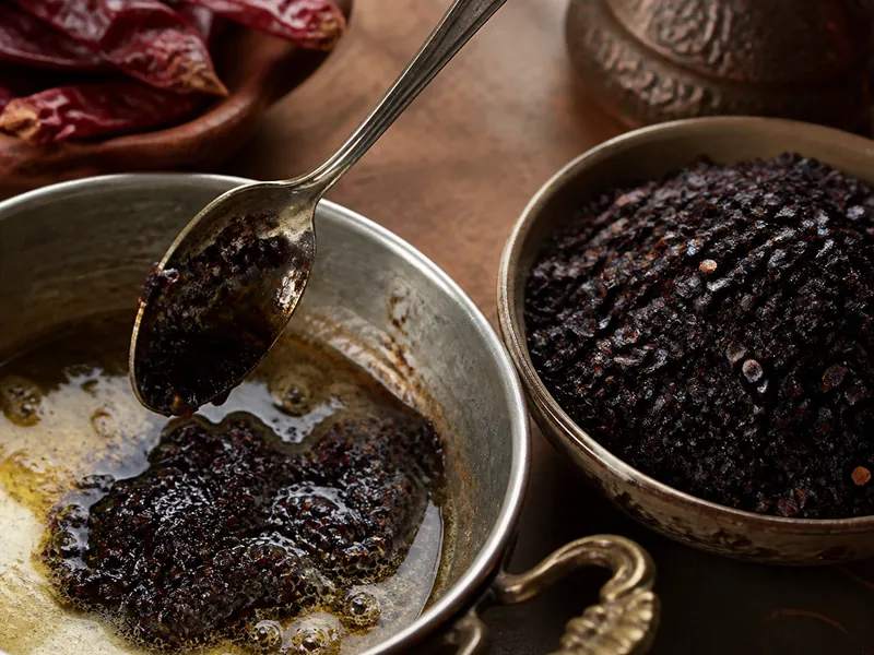 Dark purple-black urfa biber flakes with a visibly oily texture being stirred into melted butter, beside a bowl of the dry flakes