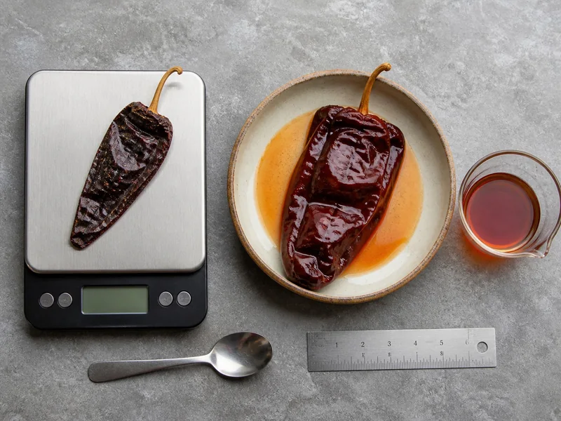 Dried chile on a gram scale beside a rehydrated chile and separate cup of soaking liquid